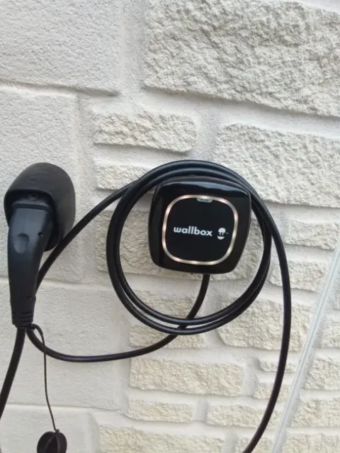 Borne de recharge murale noire avec son câble Type 2 lové sur son support, fixée sur un mur en pierre